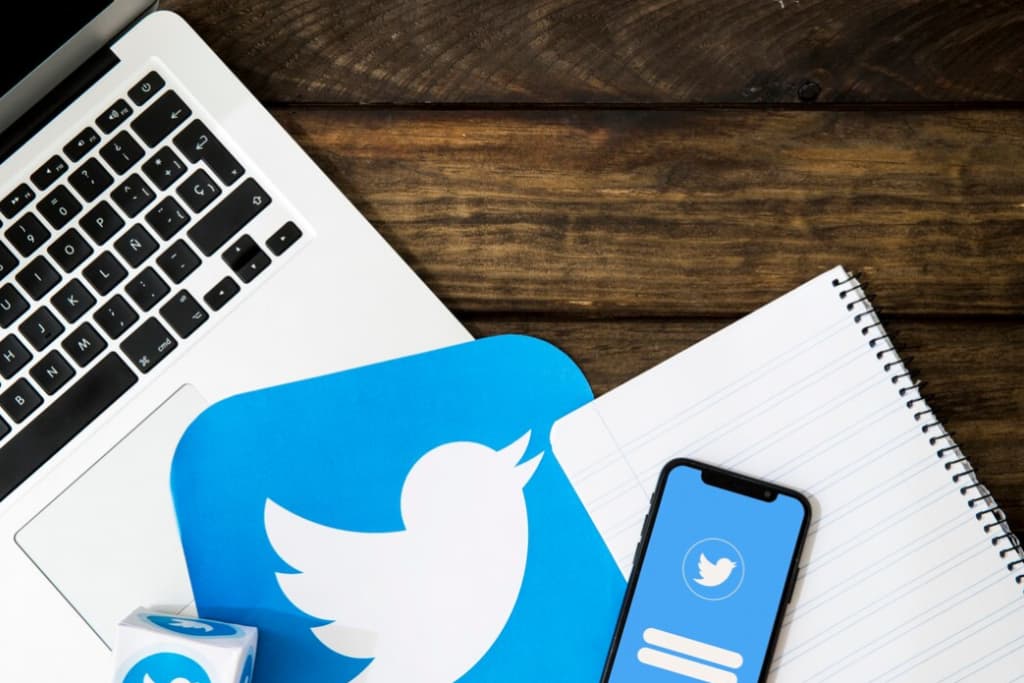 A workspace setup with a laptop, Twitter icon, notebook, and smartphone on a wooden surface